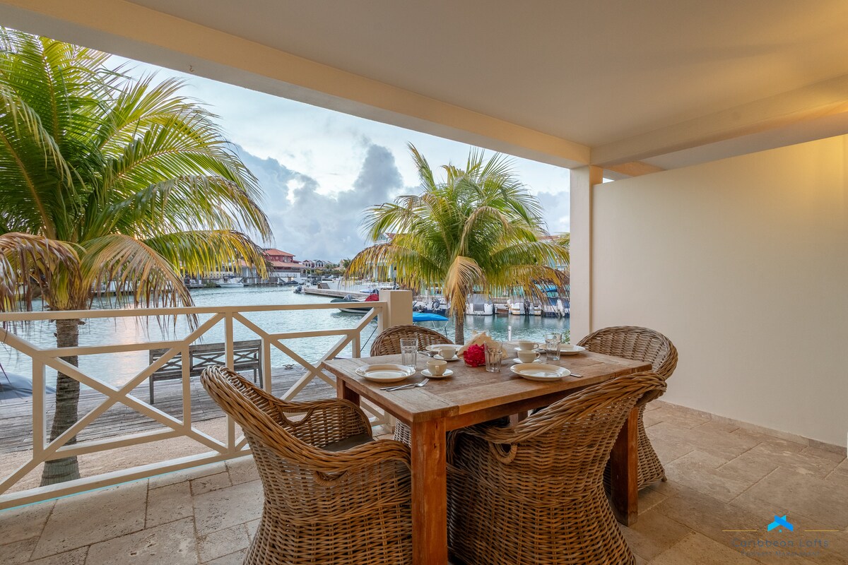 An outdoor terrace with a wooden dining table surrounded by woven chairs offers a view of palm trees and water. The table is set for a meal, with dishes, cutlery, and a flower centerpiece arranged neatly.