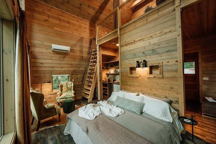 King bed, luxe bedding and pillows, wingback armchairs, sitting area, loft ladder, wall-mounted AC unit, built-in shelving, shiplap walls, vaulted wood ceiling, dark hardwood floors. Open-plan cabin bedroom.

Photo credit: Dirt and Glass