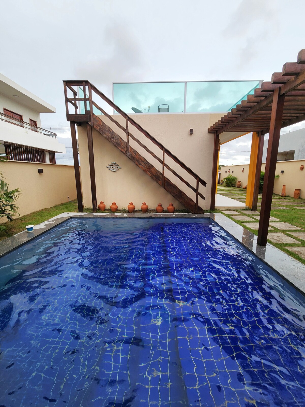 A private swimming pool is surrounded by greenery, with a wooden staircase leading to an upper terrace. The pool features a tiled blue surface, and clay pots line the back wall, adding a touch of character to the outdoor space.