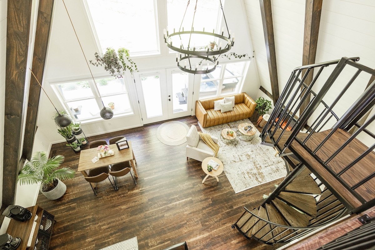 The open-concept living area features tall ceilings with exposed beams and large windows that invite natural light. A cozy brown sofa and stylish chairs surround a modern coffee table. The dining area and spiral staircase are visible, adding to the spacious atmosphere.