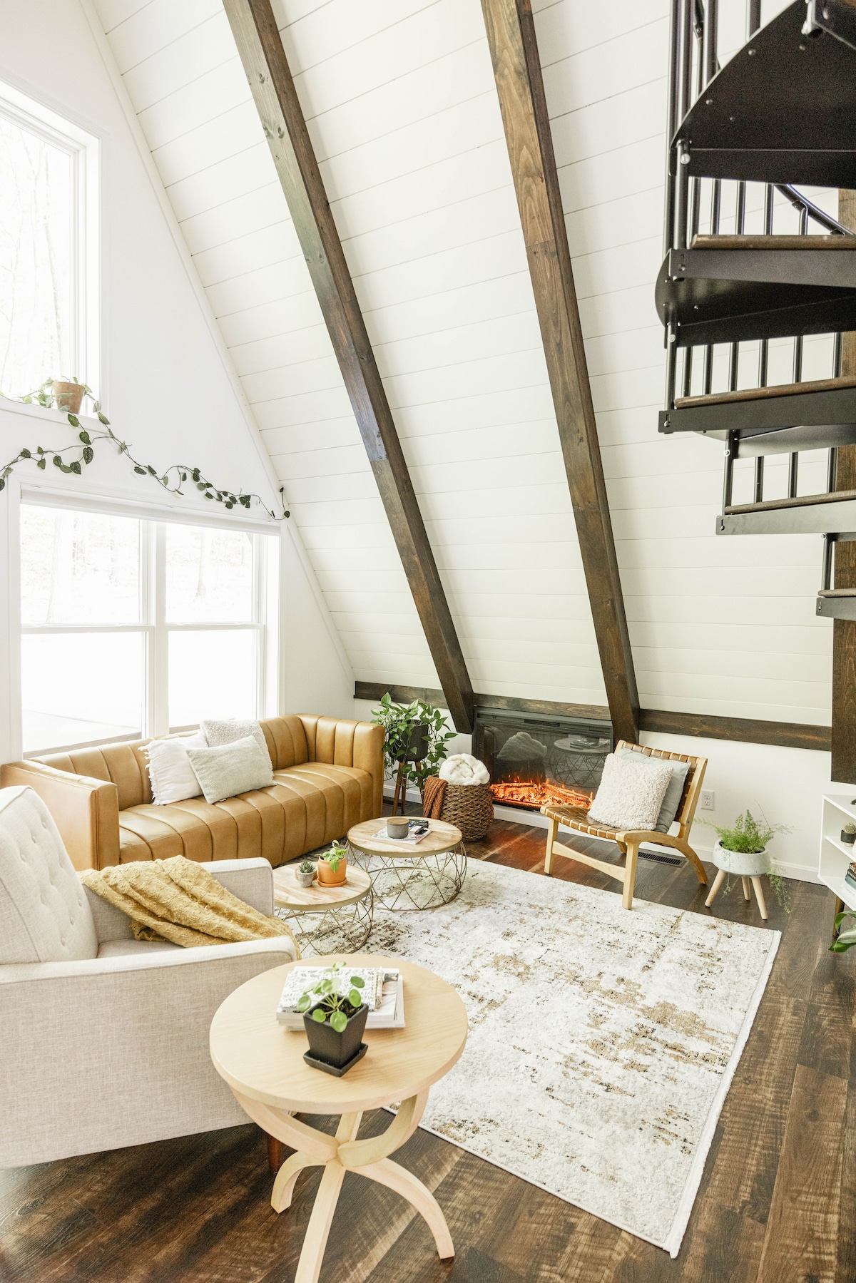 An open-concept living area is characterized by tall vaulted ceilings and large windows that allow natural light to fill the space. A plush tan sofa and stylish chairs are arranged around a decorative coffee table, near a fireplace, creating a cozy ambiance.