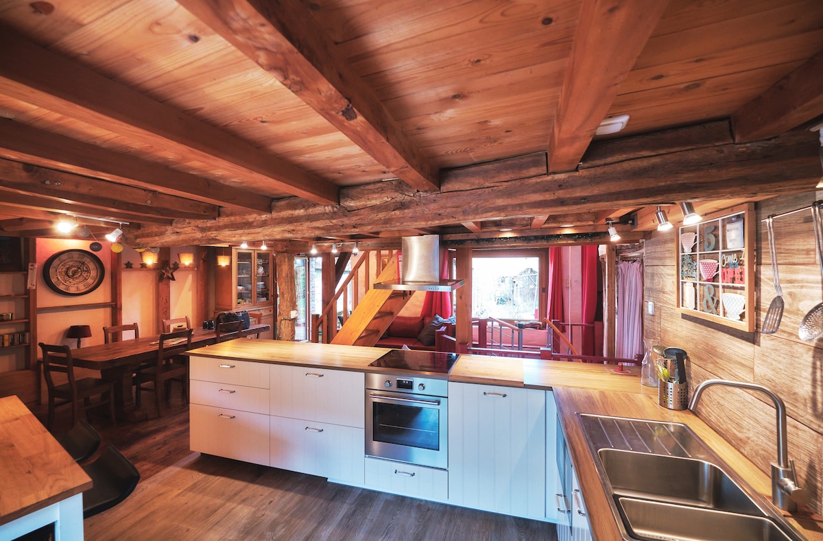 The open-plan kitchen features wooden beams on the ceiling, enhancing rustic charm. A modern kitchen island with a cooktop is visible, accompanied by a sink and ample counter space. A dining area is situated nearby, with a wooden table and chairs, bathed in natural light.