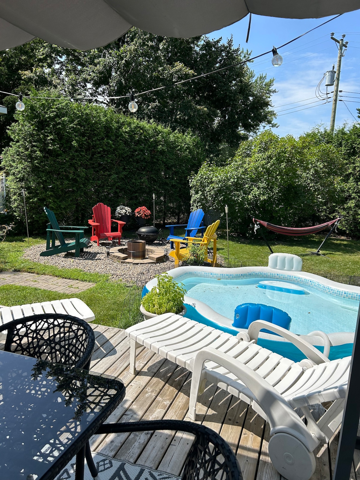 A well-kept outdoor area features a small, inviting pool surrounded by lounge chairs. Colorful Adirondack chairs in red, blue, and yellow are arranged on the grass, alongside decorative potted plants. A hammock is visible, enhancing the relaxation potential of the space.