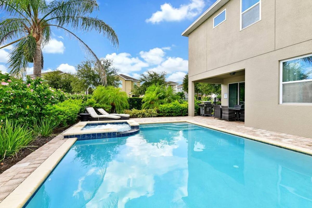 A private pool area is surrounded by lush greenery, with lounging chairs positioned beside the water. A hot tub is integrated into the pool's structure, and the home features large windows that add to the openness of the outdoor space.