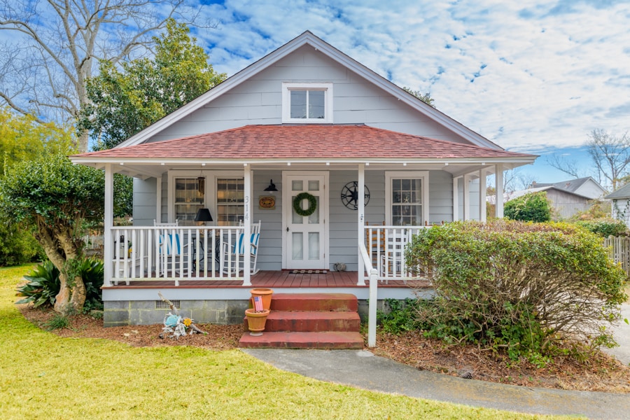 The exterior of a classic 1940s cottage is displayed, featuring a welcoming front porch with two chairs. A green wreath adorns the front door, while landscaping includes shrubs and grassy areas. The sky is partially cloudy, contributing to a bright atmosphere.