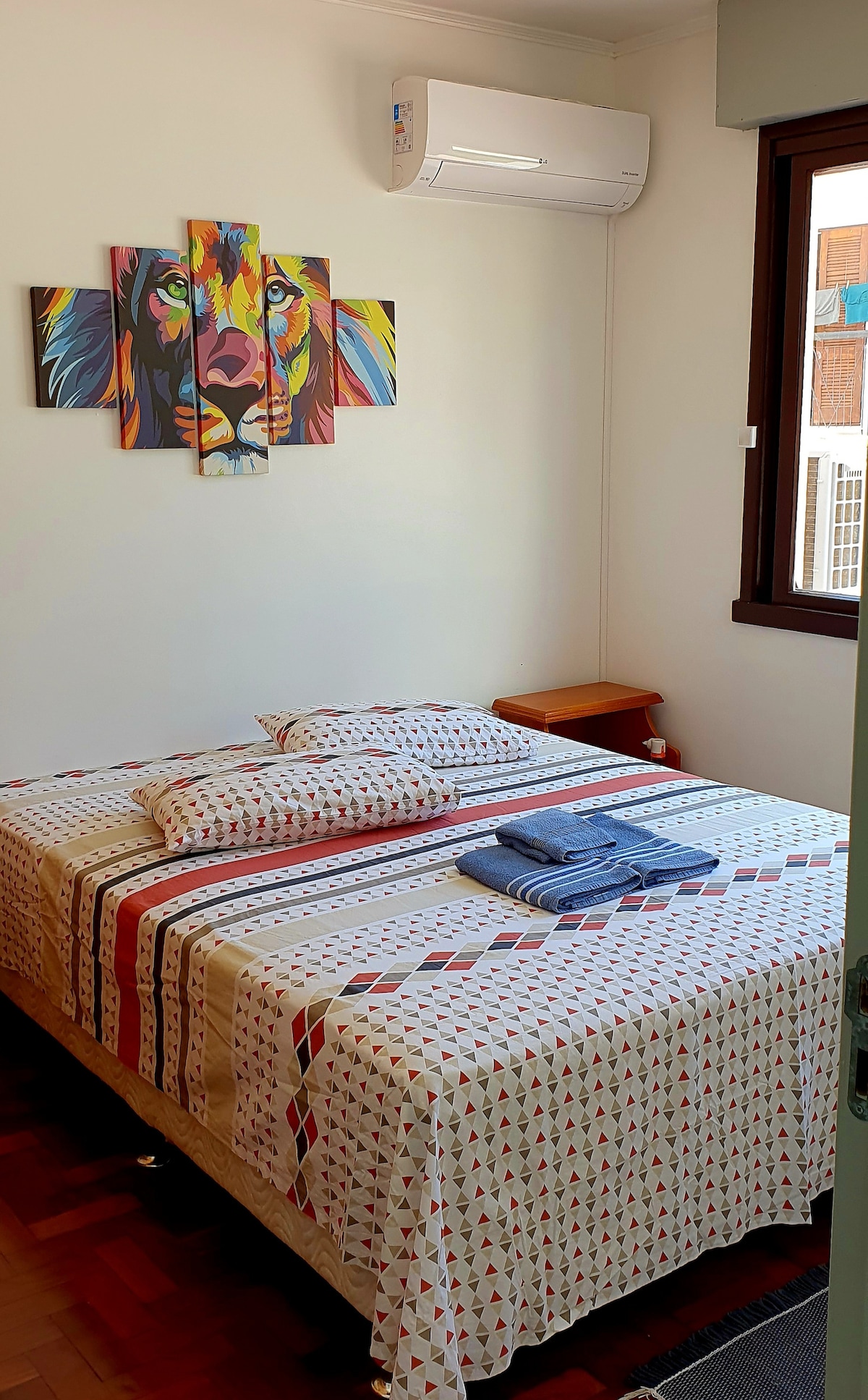 A cozy bedroom features a large bed adorned with patterned bedding and accent pillows. A vibrant lion artwork is displayed above the bed. A window brings natural light into the space, while an air conditioning unit ensures comfort.