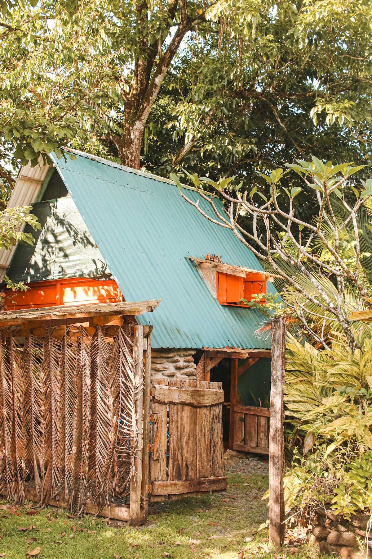 A rustic cabin with a slanted blue-green roof is surrounded by lush vegetation and fruit trees. The wooden fence features intricate weaving patterns, while vibrant orange window boxes add a pop of color against the natural backdrop.