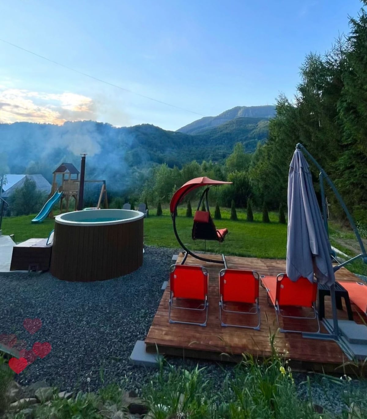 An outdoor space is shown with a round hot tub surrounded by greenery and mountain views. Red lounge chairs are arranged on a wooden deck, complemented by a swinging chair with a red canopy. A playground is visible in the background, adding to the recreational atmosphere.