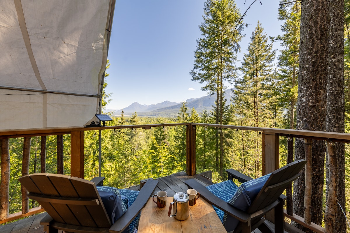 A shaded outdoor deck offers a scenic view of surrounding trees and distant mountains. Two comfortable chairs are positioned at a wooden table, which holds a teapot and cups. The peaceful setting invites guests to relax and enjoy nature.