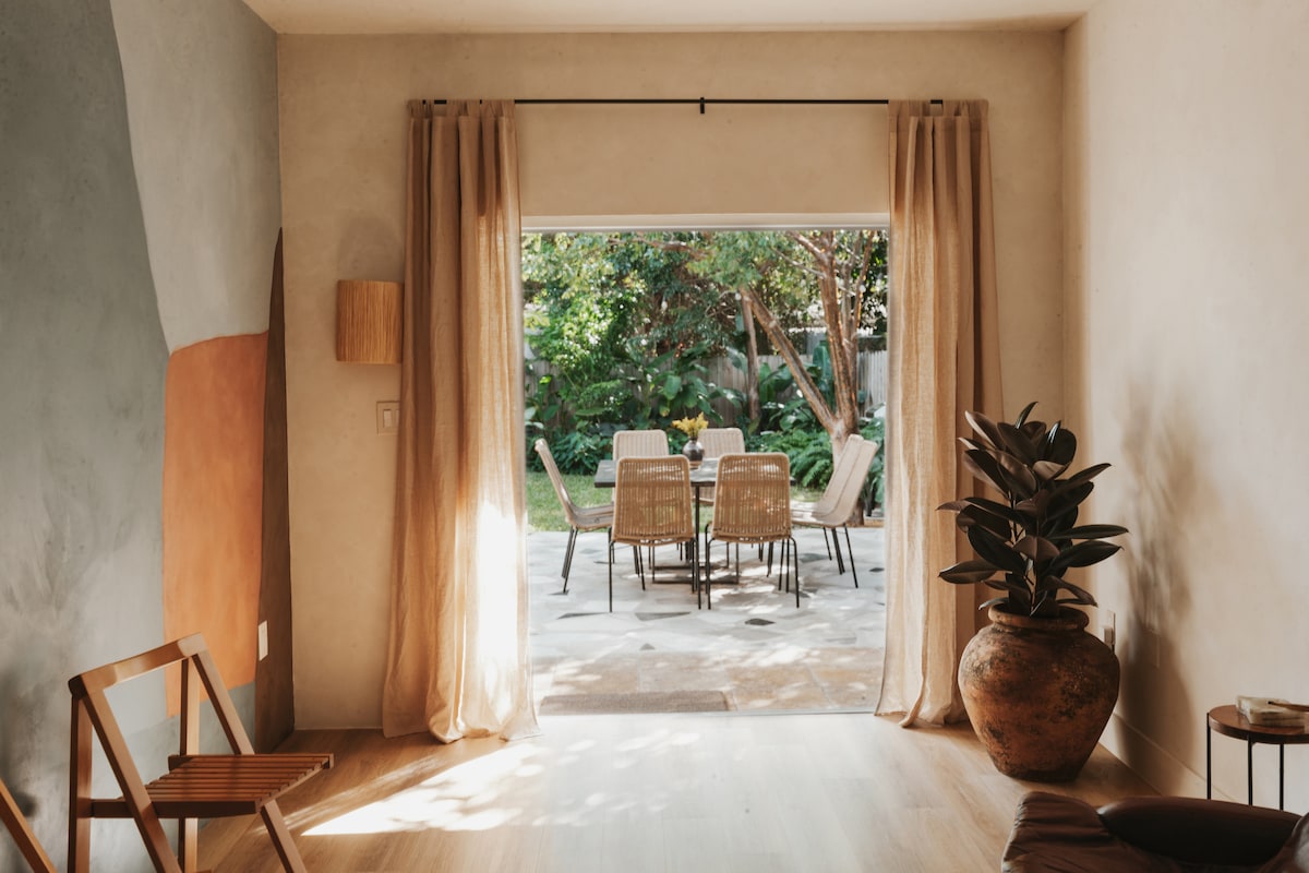 A bright indoor area opens to a private backyard, framed by light curtains. The space features a simple wooden chair, subtle wall art in earthy tones, and a potted plant. Outside, a dining table is surrounded by four chairs, set amidst lush greenery.