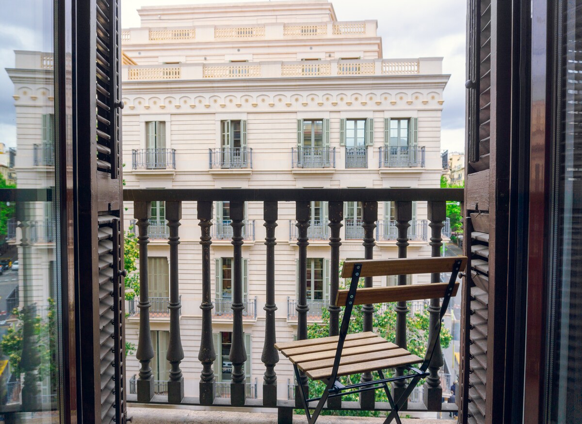 A balcony view showcases classic architecture across the street, featuring elegant façades and multiple balconies with railings. A single wooden chair is positioned on the balcony, allowing for a comfortable space to enjoy the surroundings.