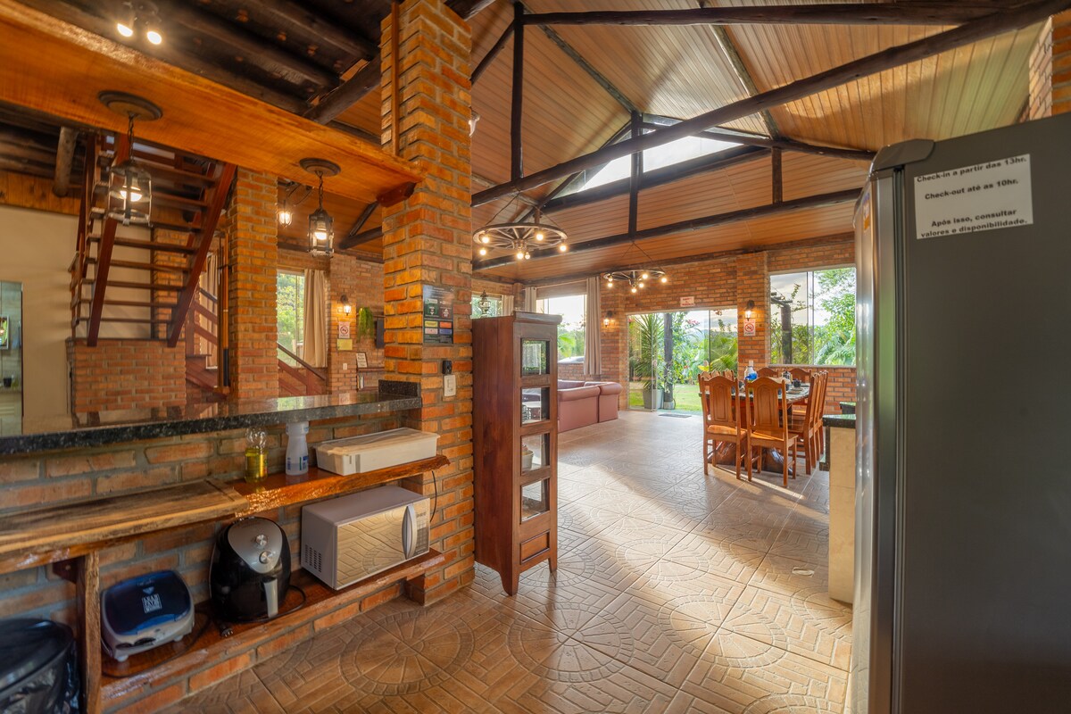 The spacious common area features a large kitchen with wooden accents and brick walls. Natural light streams in through expansive windows, showcasing a dining table suitable for multiple guests. A staircase leads to the upper level, while comfortable seating is provided nearby.