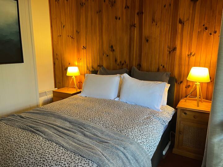 ห้องนอนหลักมีเตียงควีนไซส์ โต๊ะข้างเตียง โคมไฟข้างเตียง พัดลมเพดาน และตู้เสื้อผ้า
มองเห็นระเบียงด้านข้างและพื้นที่สวน