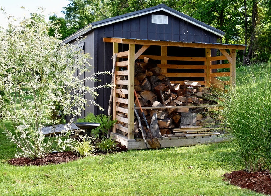 A wood shed is situated in a landscaped yard, featuring neatly stacked firewood under a wooden overhang. Surrounding greenery includes flowering shrubs and tall grasses, creating a natural setting. Fresh mulch outlines the area, contributing to the tidy appearance of the outdoor space.