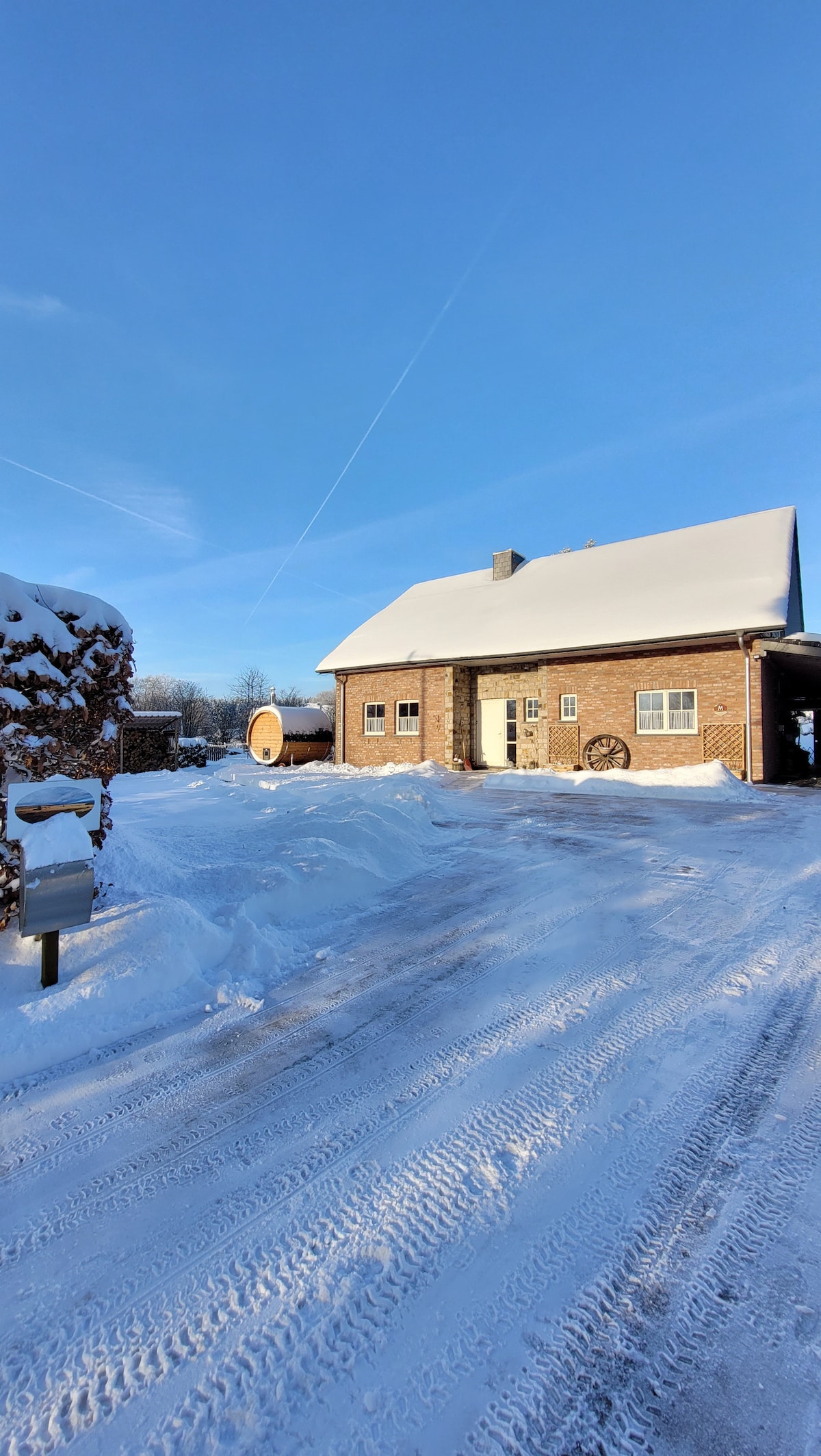 A two-story brick house is seen with a sloped roof, partially covered in snow. A pathway is visible, leading to the entrance, with snow blanketing the ground. To the side, a traditional wooden sauna can be glimpsed amidst the peaceful winter landscape.