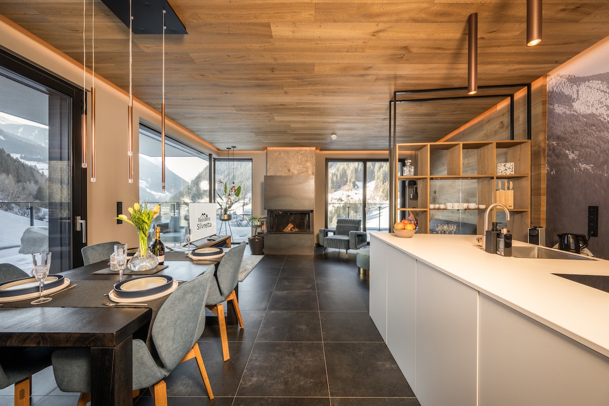 The open-plan kitchen and dining area feature a modern design with a large dining table that accommodates up to four people. A sleek kitchen island is visible, accented by elegant lighting, while a cozy seating area with a fireplace is present nearby, blending comfort and style.