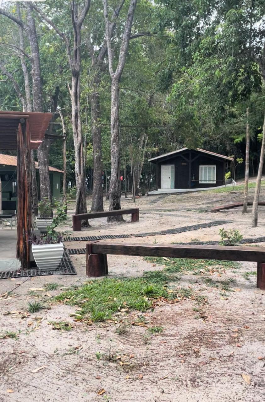 A rustic outdoor setting features a wooden bench positioned on a sandy area. Surrounded by tall trees, a cozy cabin can be seen in the background, along with additional structures showcasing simple architecture, creating a peaceful environment for relaxation.