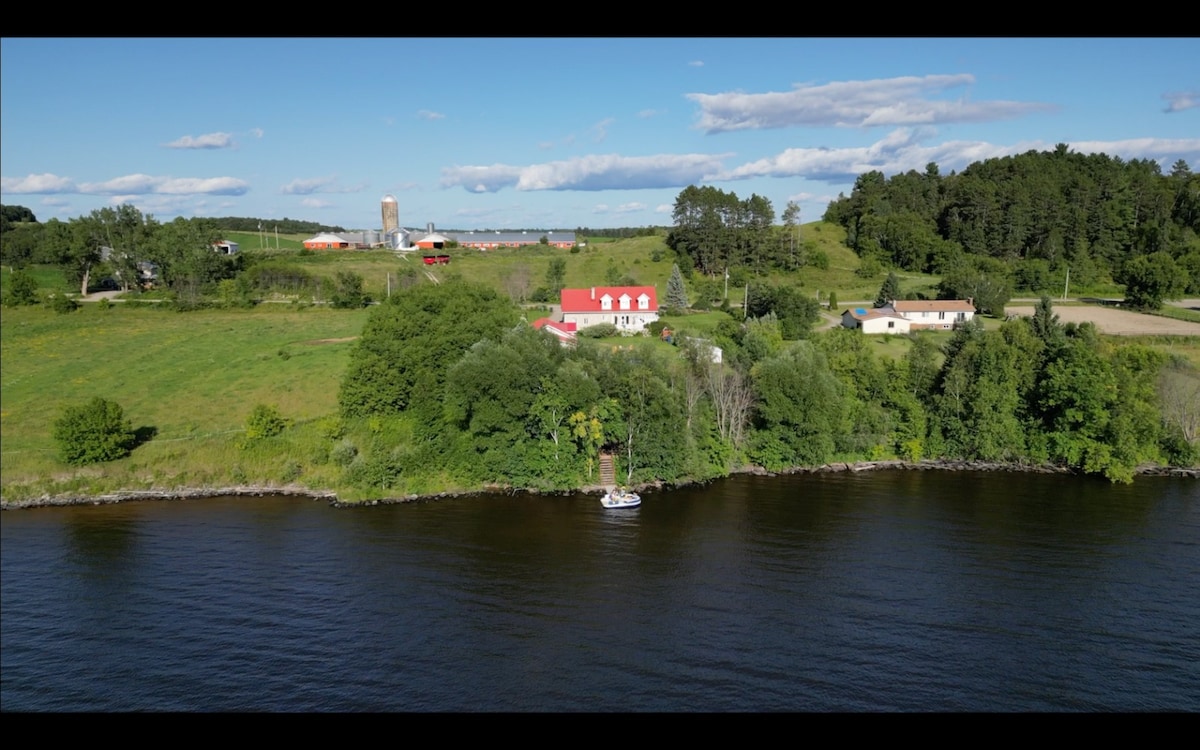 Aerial view of a private house situated along the shores of Lake Timiskaming, surrounded by lush greenery. The vibrant red roof contrasts with the blue water and is complemented by the expansive farmland in the background, emphasizing the tranquil setting.