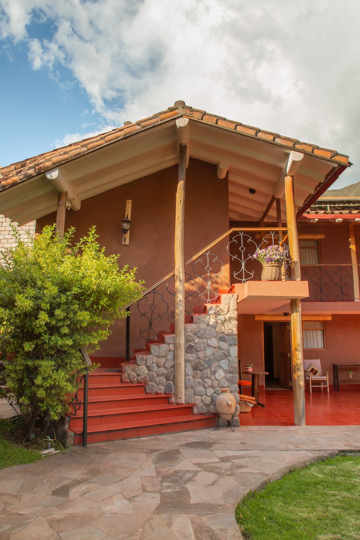 The exterior of a charming home is shown, featuring a warm terracotta façade and a tiled roof. An inviting staircase leads to a covered entrance, complemented by a stone accent wall. Lush greenery and decorative planters enhance the surrounding area.