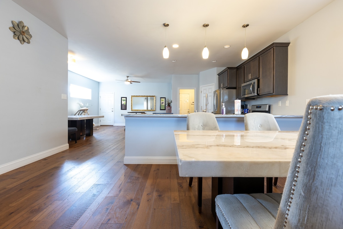 An open dining area showcases a marble-topped table surrounded by upholstered chairs, with warm wood flooring extending throughout. The well-lit kitchen features modern cabinetry and stainless steel appliances, while a spacious living area can be seen in the background.