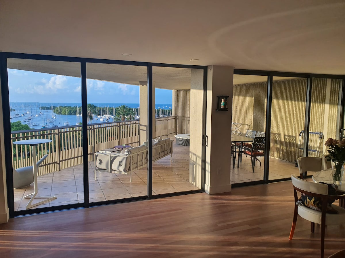 A spacious living area opens onto a large balcony, featuring panoramic views of the marina. Floor-to-ceiling glass doors invite natural light into the room. A modern dining set and comfortable seating arrangements are visible, complemented by a warm wooden floor.