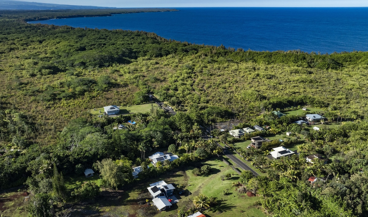 An aerial view showcases a lush green landscape bordered by the blue coastline. Various houses are positioned throughout the area, with roads winding through the greenery. The ocean shimmers in the distance, complementing the vibrant natural surroundings.