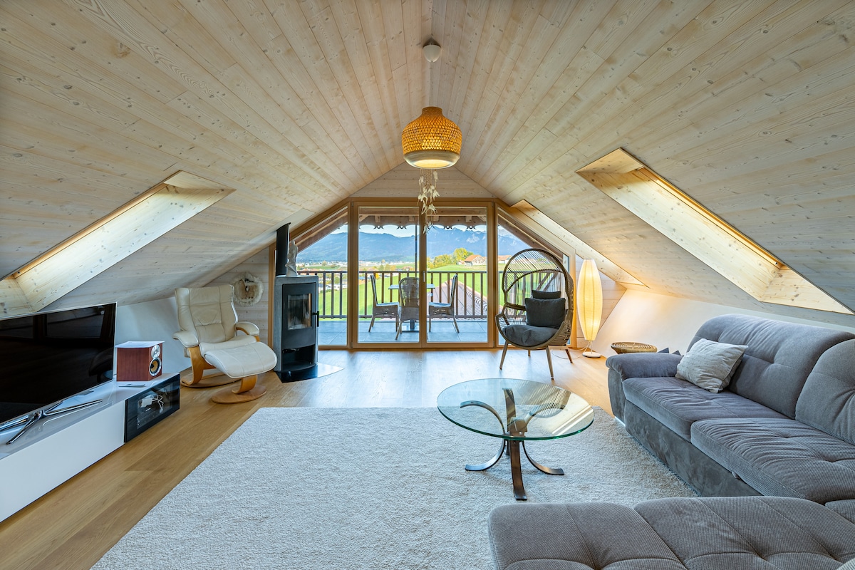 An open loft space features a sloped wooden ceiling, a large glass door leading to a balcony with a view. A comfortable sectional sofa and a stylish armchair are complemented by a glass coffee table. Natural light fills the area, enhancing the serene atmosphere.