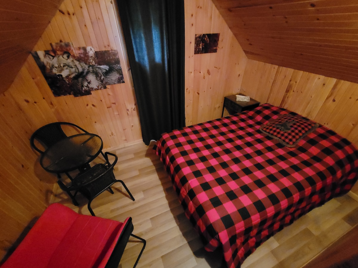 A compact sleeping area features a double bed with a red and black checkered blanket. Two small chairs and a round table occupy one corner, and wooden walls create a cozy atmosphere. Dark curtains cover a small window, enhancing privacy.