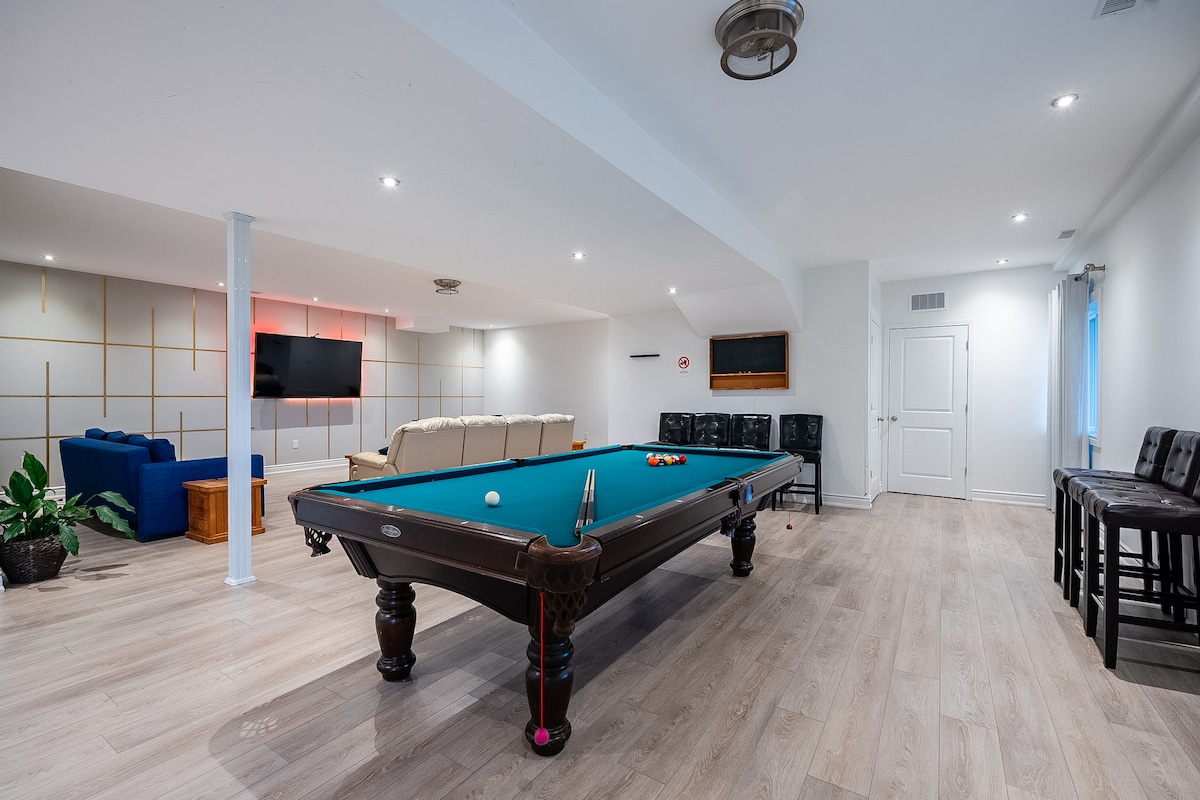 A spacious lower-level entertainment area features a billiards table with a vibrant blue surface. Comfortable seating is arranged nearby alongside a television mounted on the wall. Soft lighting creates a welcoming atmosphere, enhancing the functionality of this versatile space.