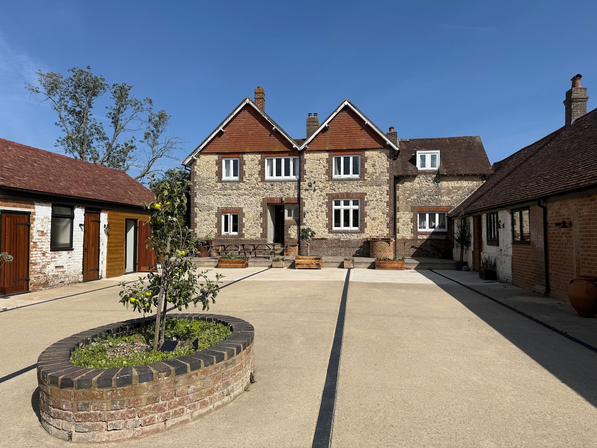 The farmhouse features a stone façade with a warm, traditional style. A spacious courtyard is paved and framed by planters. Surrounding buildings include stable conversions and storage, all set against a clear blue sky.