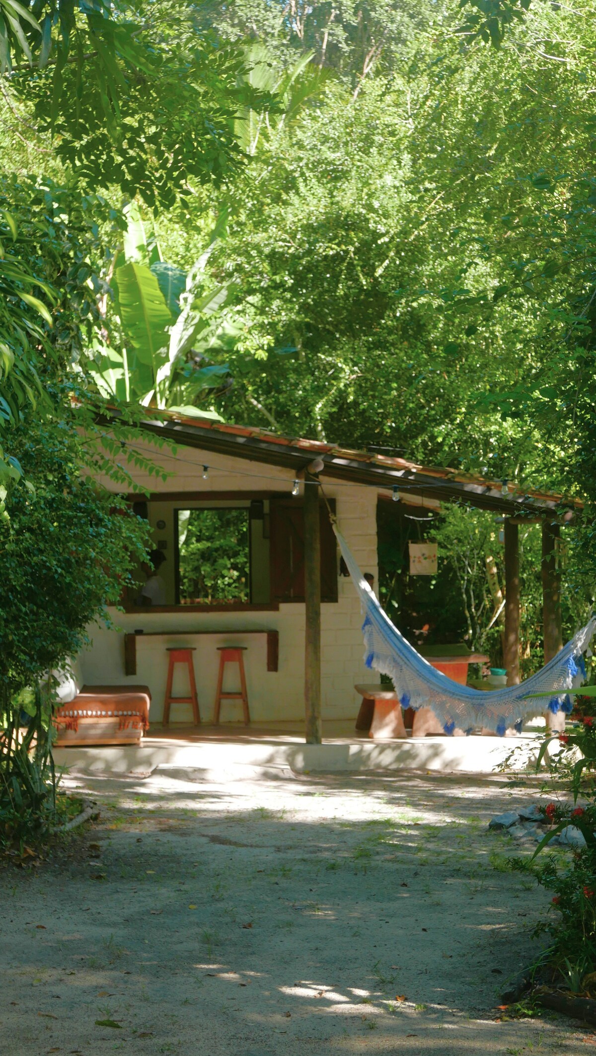 A charming cabin is nestled among lush greenery, featuring a shaded outdoor area with a bar and seating. Hammocks are suspended from the porch, inviting relaxation in the serene environment. A sandy path leads to the cabin, enhancing the connection with nature.