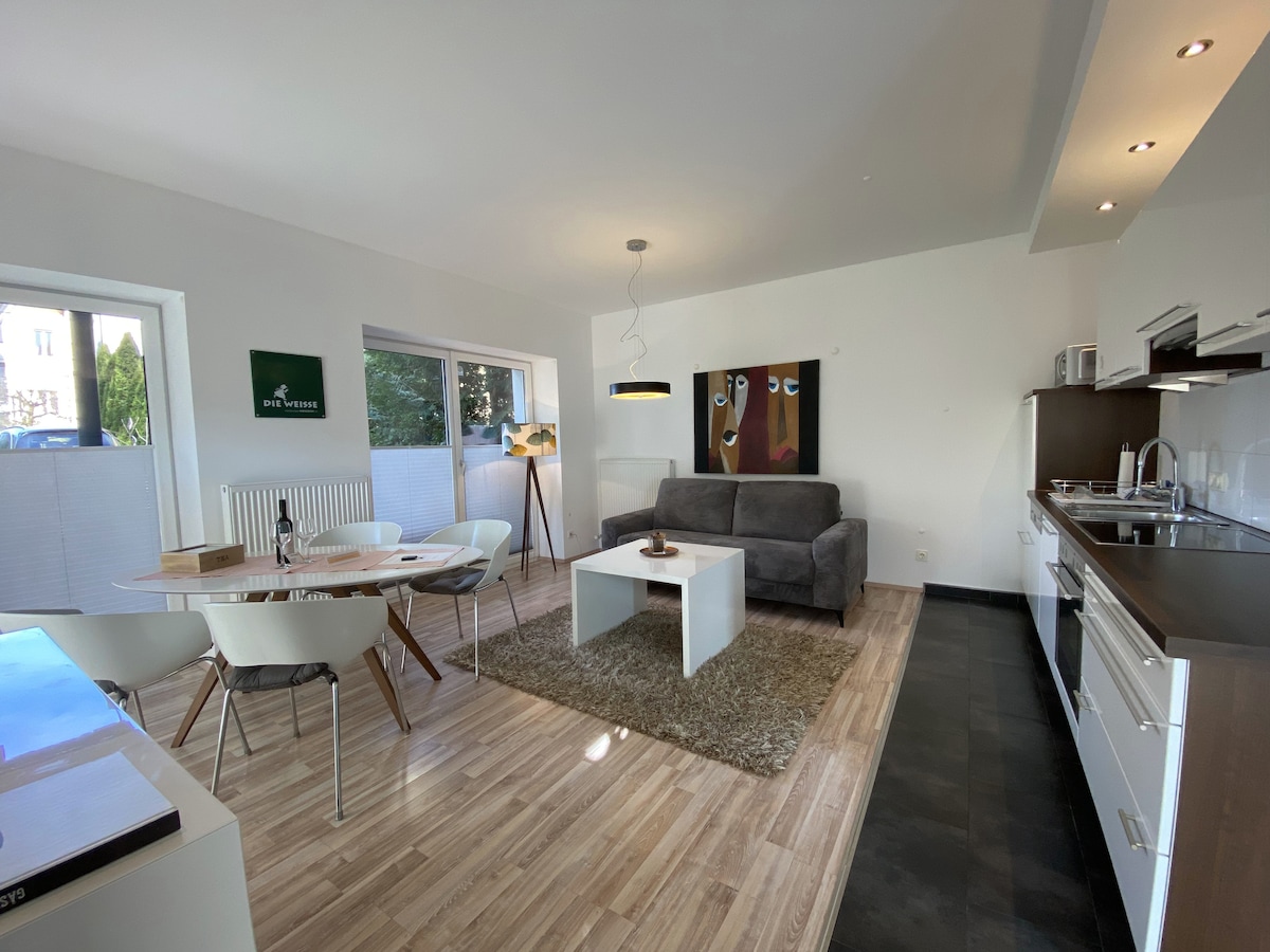 A modern living area showcases an open layout with a comfortable gray sofa facing a minimalist coffee table. A dining table with white chairs is positioned nearby. Large windows allow natural light to fill the space, enhancing the neutral color palette of the interior.