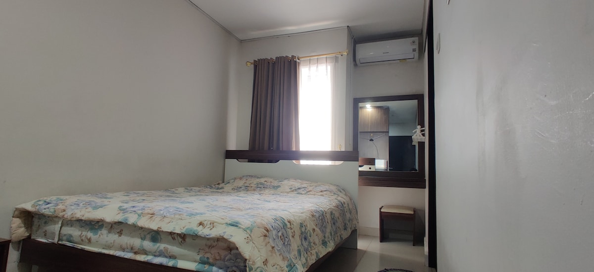 A well-appointed sleeping area is shown, featuring a spacious bed with a floral-patterned blanket. Natural light enters through a window covered by light curtains. A large mirror and a small stool are positioned nearby, with warm-toned curtains framing the window.