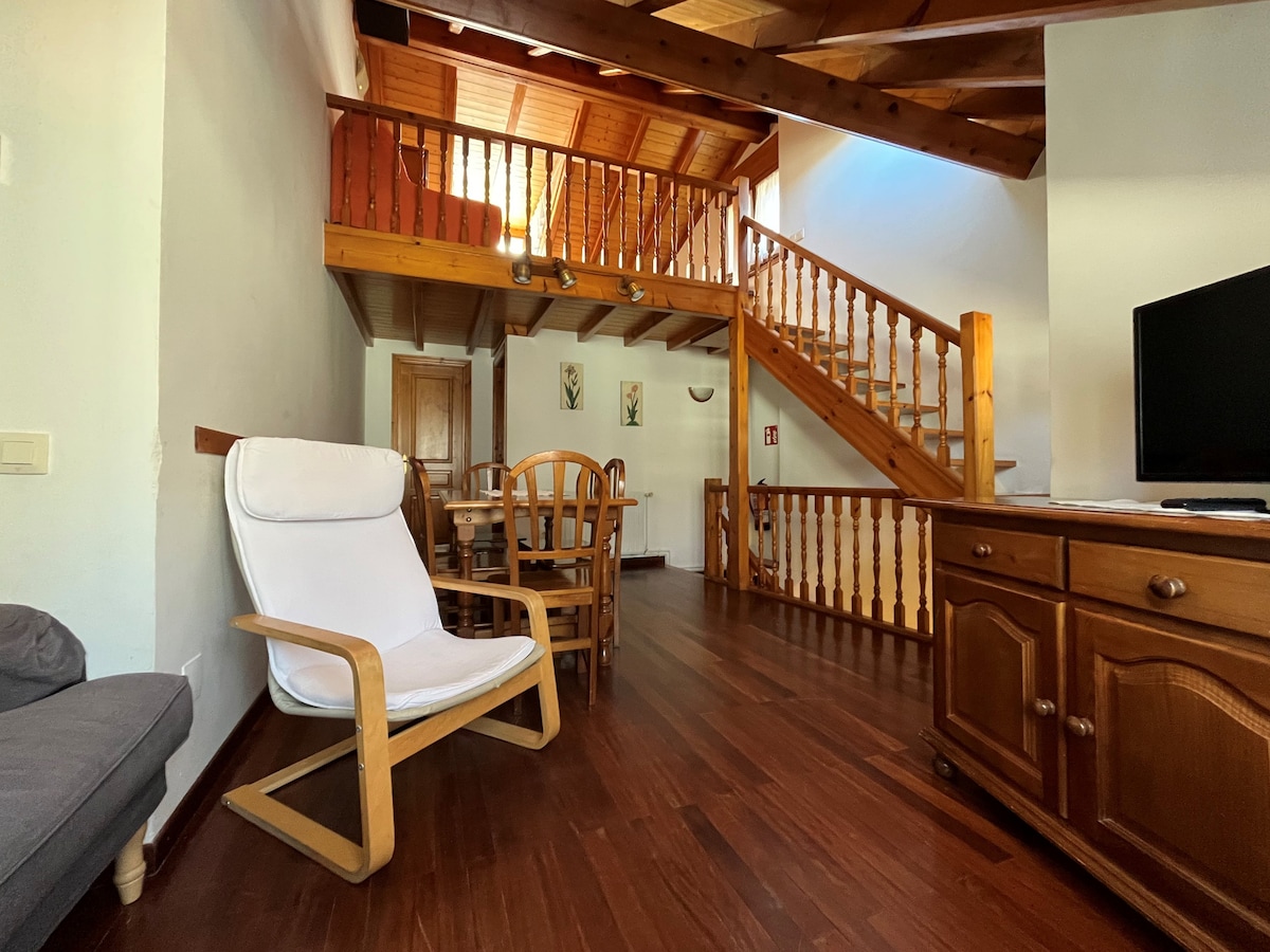 A warm wooden staircase leads to the upper level, while a cozy seating area features a light armchair and a grey couch. A dining table can be seen in the background, surrounded by wooden chairs, with natural light illuminating the space.