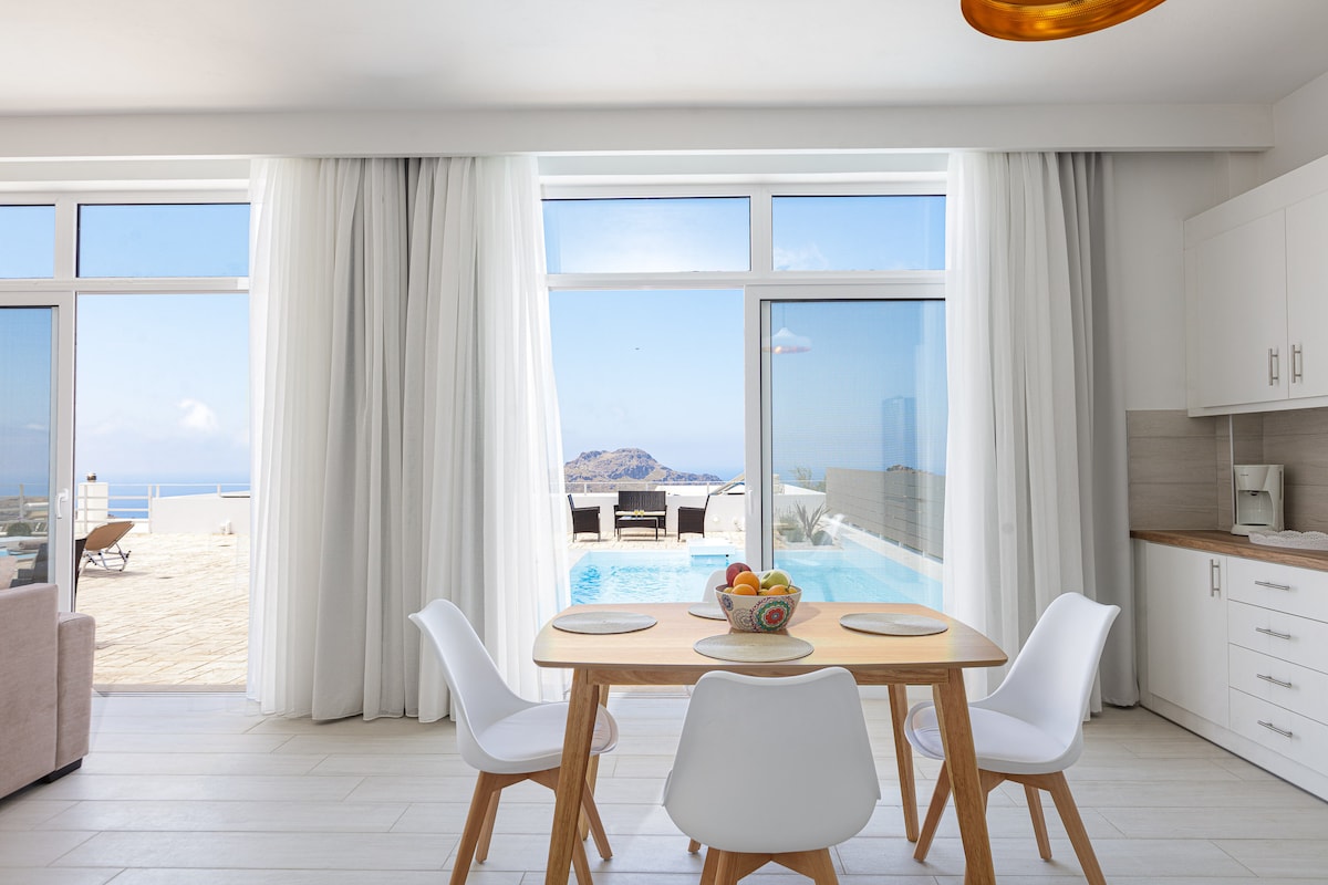 A dining area features a wooden table surrounded by minimalist white chairs. Large glass doors allow natural light to fill the space while providing a view of the outdoor pool and scenic landscape beyond. Light, sheer curtains elegantly frame the windows.