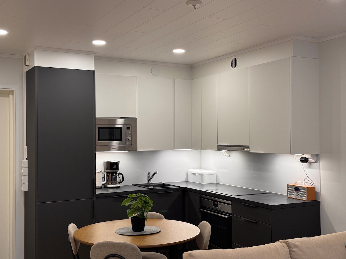 A modern kitchen features sleek cabinetry in black and white. Essential appliances, including a microwave, coffee maker, and dishwasher, are visible. A round dining table, accompanied by four chairs, is placed near a small potted plant, creating a practical and functional dining space.