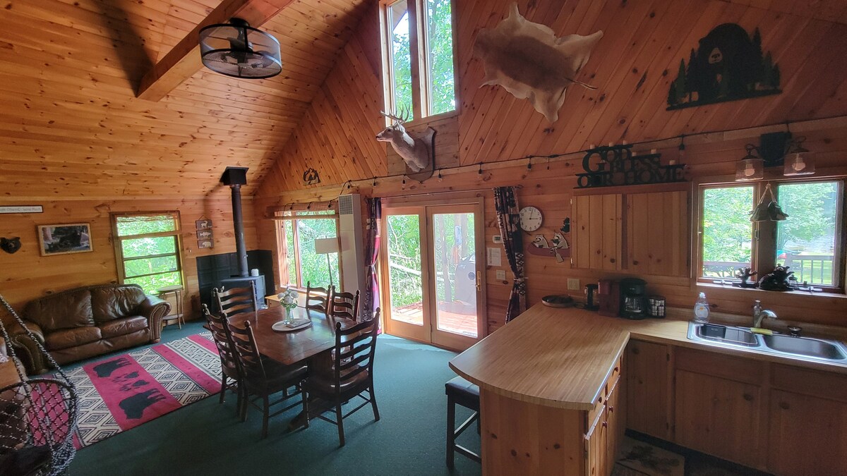 The open living area showcases a wooden ceiling, large windows, and a cozy seating arrangement with a brown sofa. A dining table with multiple chairs is positioned near the kitchen, which features a wooden countertop. Natural light fills the space, enhancing the cabin's inviting ambiance.
