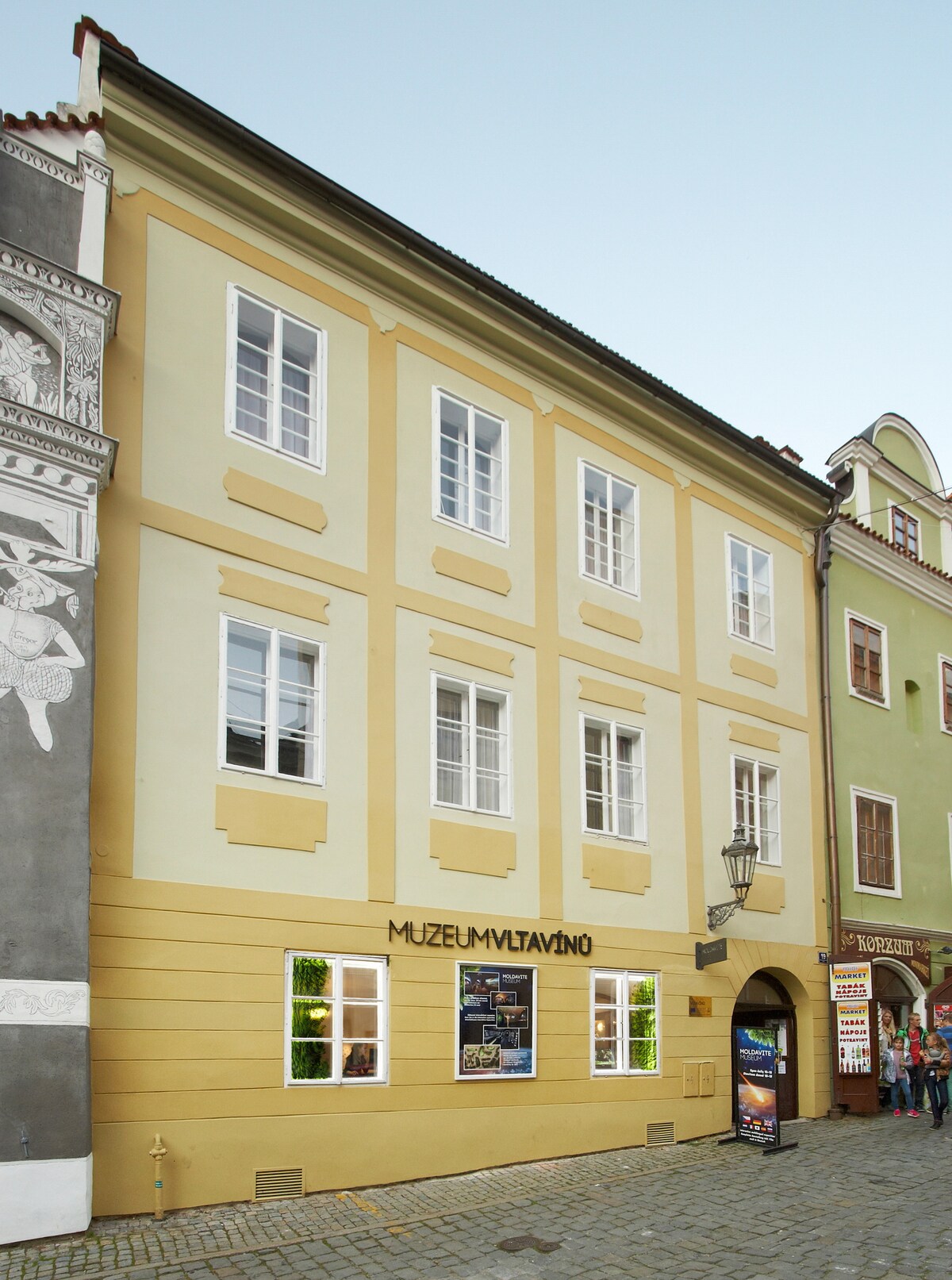 The exterior of the Vltavín Museum features a two-story yellow building with decorative elements and multiple windows. A sign displaying 'MUZEUM VLTAVÍNŮ' is prominently placed on the front. The adjacent architecture reflects a historic charm, enhancing the cultural surroundings.