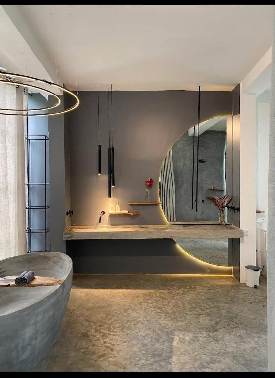 A modern bathroom is featured, highlighting a sleek, curved sink atop a wooden counter. Soft lighting outlines a rounded mirror, and a small vase with a red flower adds a touch of color. The space combines concrete and smooth surfaces for a sophisticated feel.