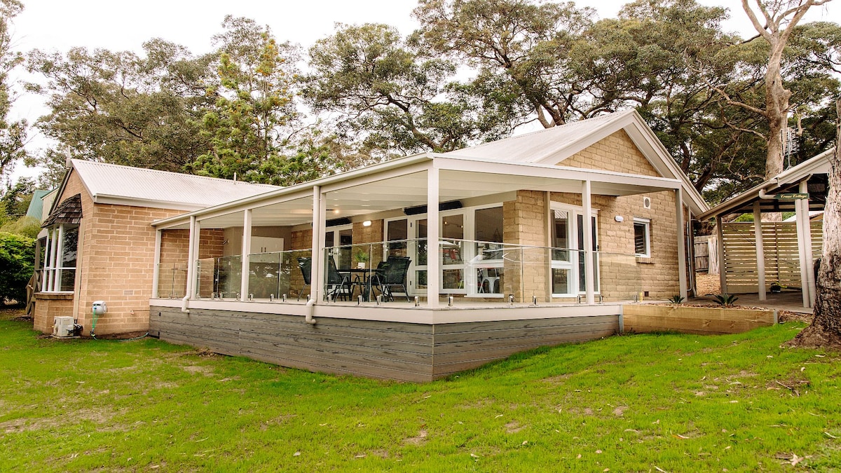 The cottage features a contemporary exterior with stone and wooden elements. A covered deck wraps around the front, providing space for outdoor seating. Lush green grass surrounds the building, while trees provide natural shade. Large glass windows allow for abundant natural light inside.