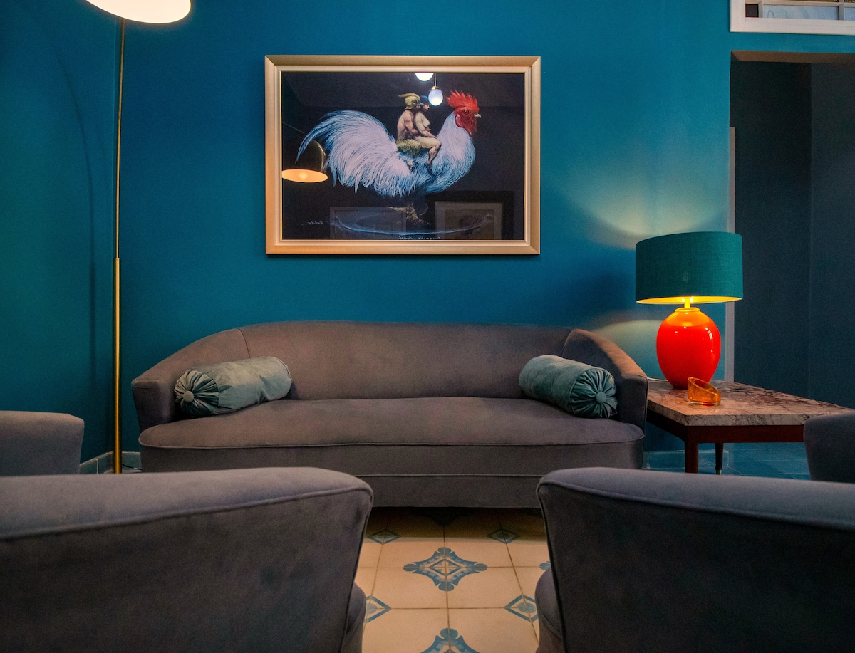 A cozy seating area features a curved sofa in a soft grey tone, complemented by two armchairs. A colorful artwork depicting a rooster hangs above the sofa. An orange lamp and a small table can be seen, adding to the room's vibrant decor.