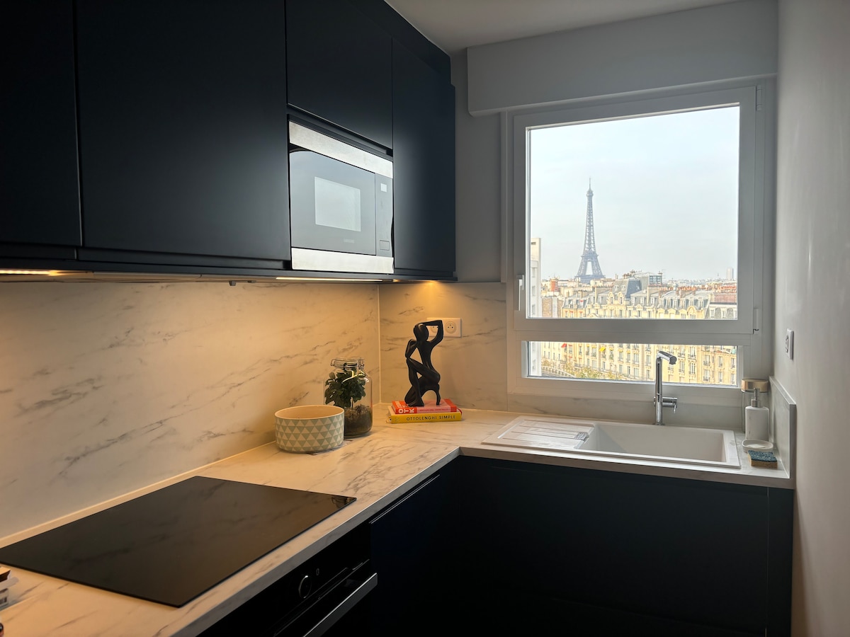 The kitchen features dark cabinetry and a marble countertop, providing a modern aesthetic. A window frames a view of the Eiffel Tower and the Paris skyline. Visible are a sink, stove, and decorative sculpture, creating a functional and visually appealing cooking space.