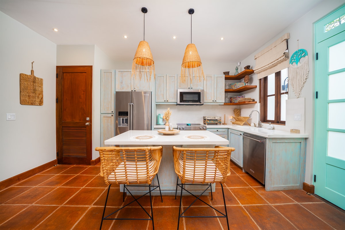 A well-equipped kitchen features light wood cabinetry and stainless steel appliances. A central island with seating for two is adorned with a decorative centerpiece. Natural light filters through the window, creating a bright and airy cooking space accented by woven pendant lights.