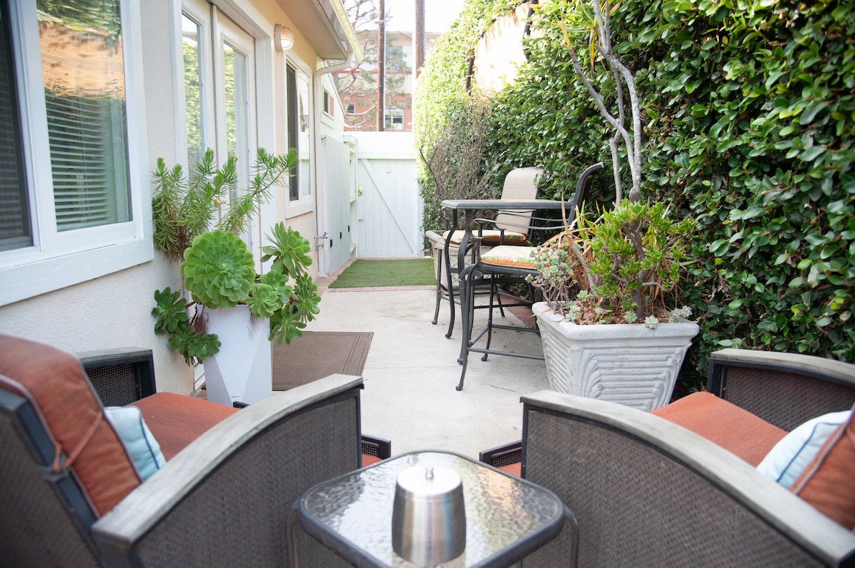 A serene outdoor patio area is depicted, featuring a small table surrounded by four chairs. Comfortable seating is arranged with cushions, and decorative pots filled with greenery add a touch of nature. The space is enclosed by lush greenery, providing a sense of privacy.