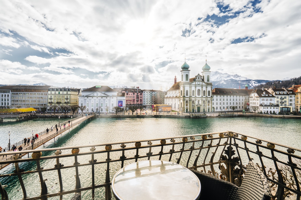 A balcony view showcases the shimmering Reuss River and the iconic Chapel Bridge with its water tower. The picturesque buildings lining the waterfront and distant mountains are visible under a wide, cloud-streaked sky, creating a serene atmosphere.