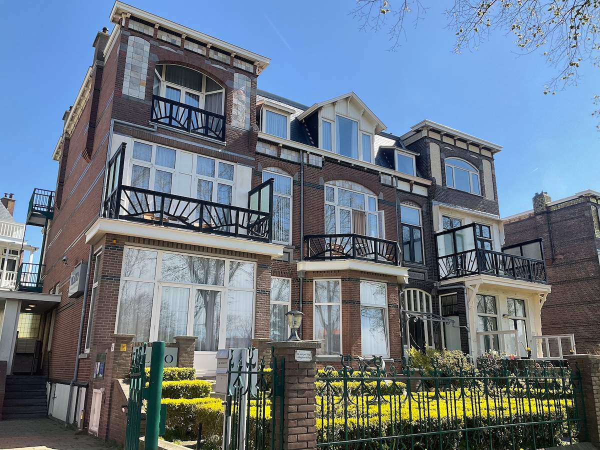 The classical building features a red brick facade with multiple balconies adorned with black railings. Large windows allow natural light to fill the interior. A well-maintained garden and green hedges surround the property, contributing to the tranquil atmosphere of the location.