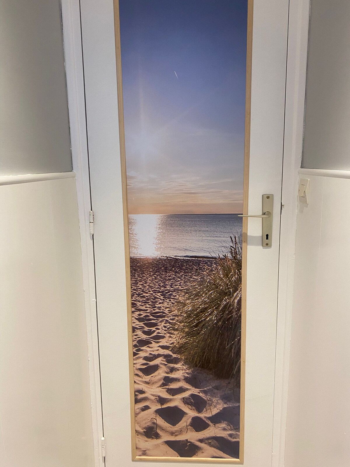 A door features a large image of a sandy beach, with tall grasses framing the scene. Gentle waves can be seen lapping at the shore under a clear sky, illuminated by the soft light of the setting sun.