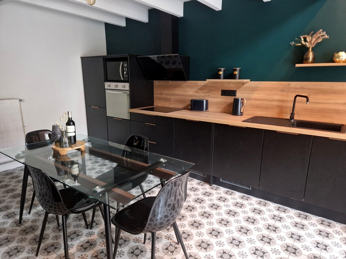 The kitchen area features modern cabinetry in a sleek black finish with wood accents. A glass dining table with four black chairs is positioned centrally, while appliances such as an oven and kettle are neatly arranged along the countertop. Light fixtures above add a warm glow to the space.