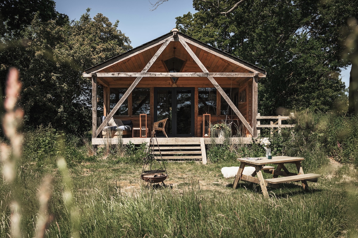 A rustic cabin is set in a natural landscape, featuring a wooden deck with chairs facing large glass doors. A picnic table and a fire pit are visible in the foreground, surrounded by tall grass and greenery, adding to the tranquil outdoor setting.