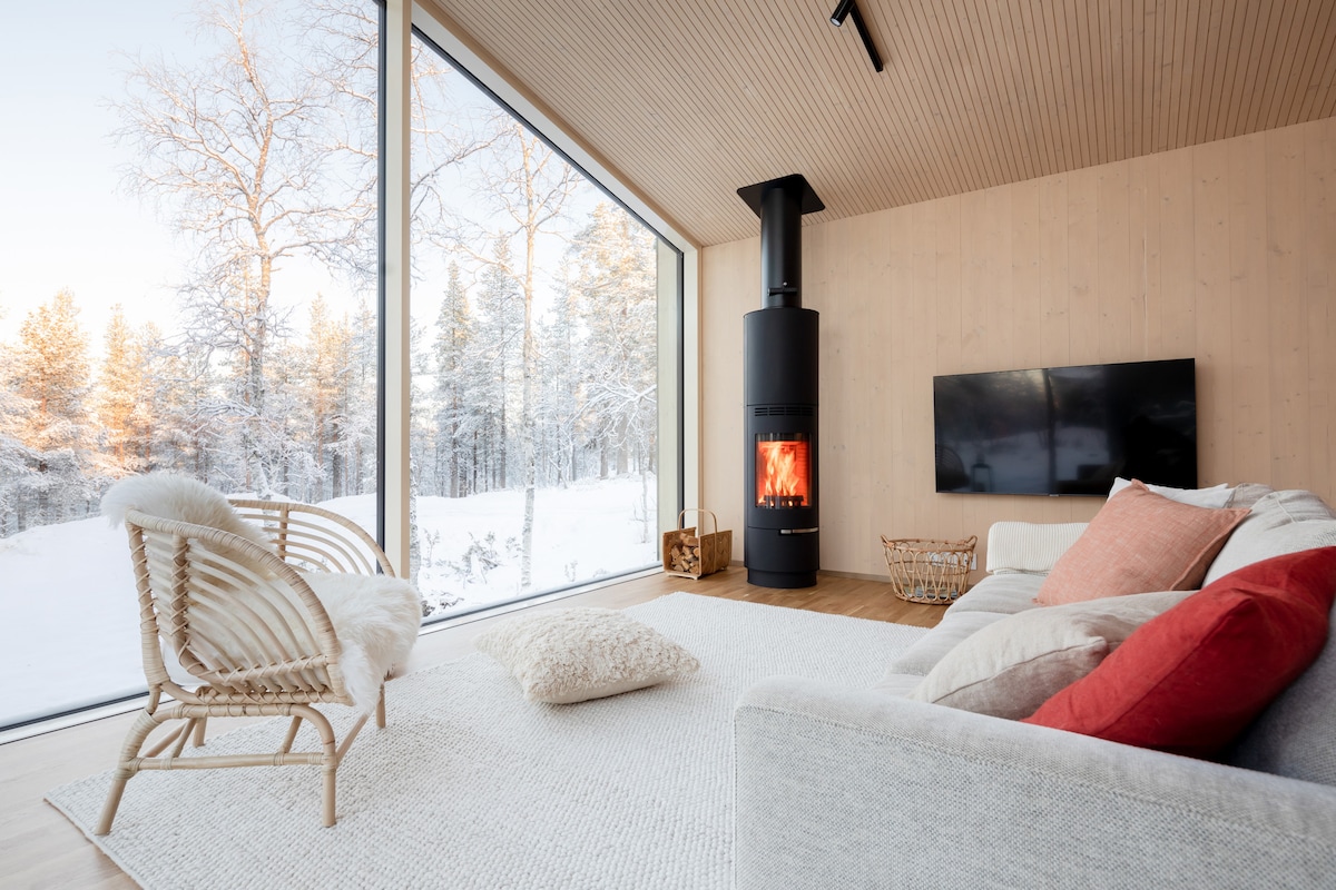 A spacious living area is highlighted by large landscape windows that offer unobstructed views of the snowy outdoors. A black fire stove stands beside a contemporary TV mounted on the wall. Comfortable seating options include a light-colored sofa and woven chairs, along with a soft area rug.